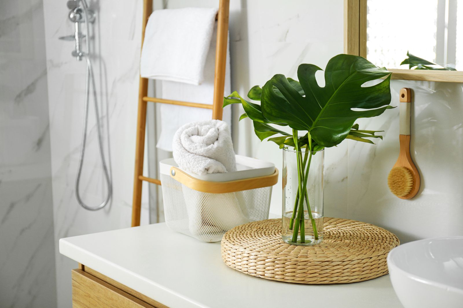 Waschtisch mit Drahtkorb und gerolltem Handtuch, daneben Monstera-Blätter in einer Glasvase auf einem Rattan-Untersetzer, dahinter eine Handtuchleiter