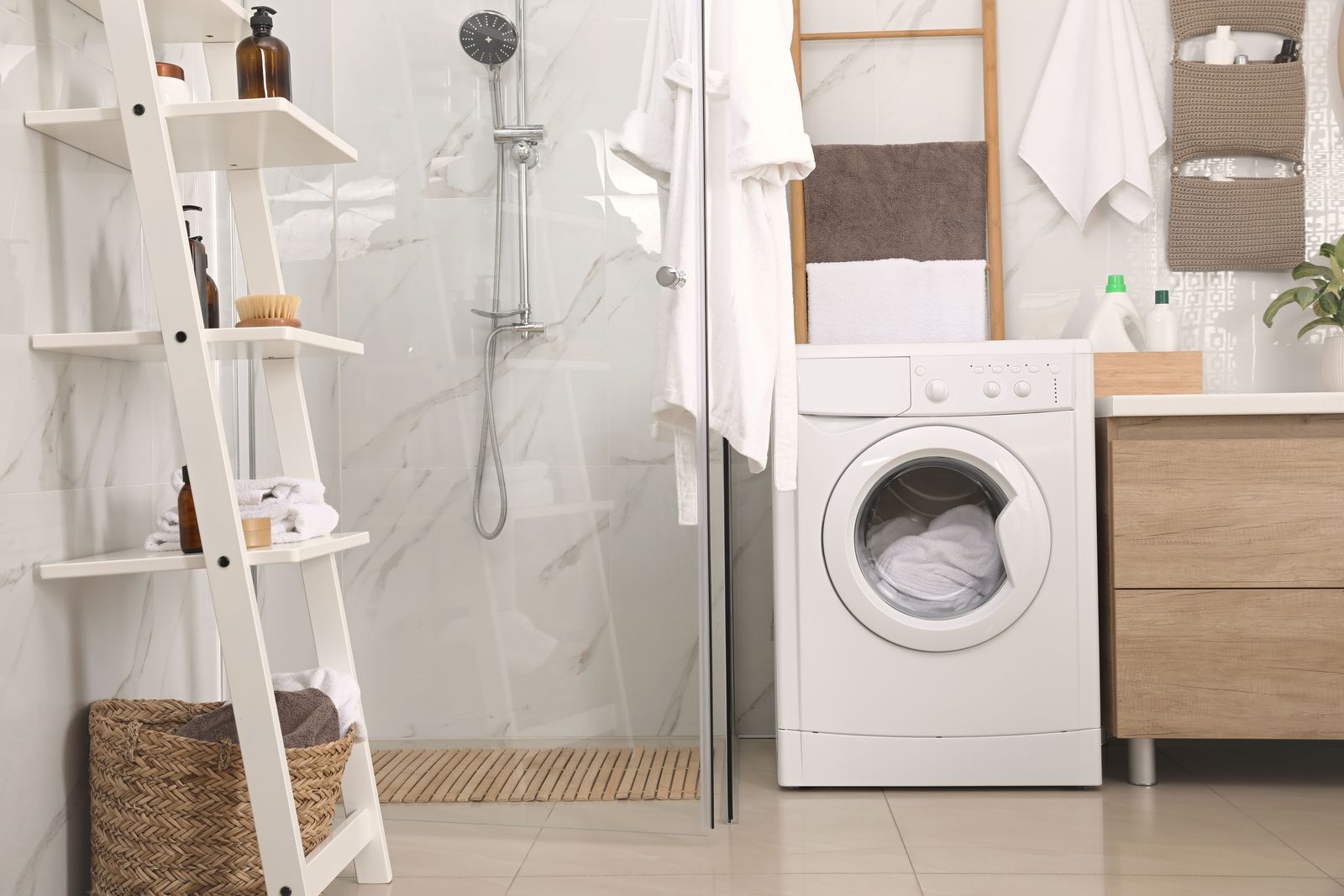 Helles Badezimmer mit weißem Leiterregal voller Handtücher und Flaschen, Rattankorb, Glasdusche, Waschmaschine und einer Handtuchleiter aus Holz