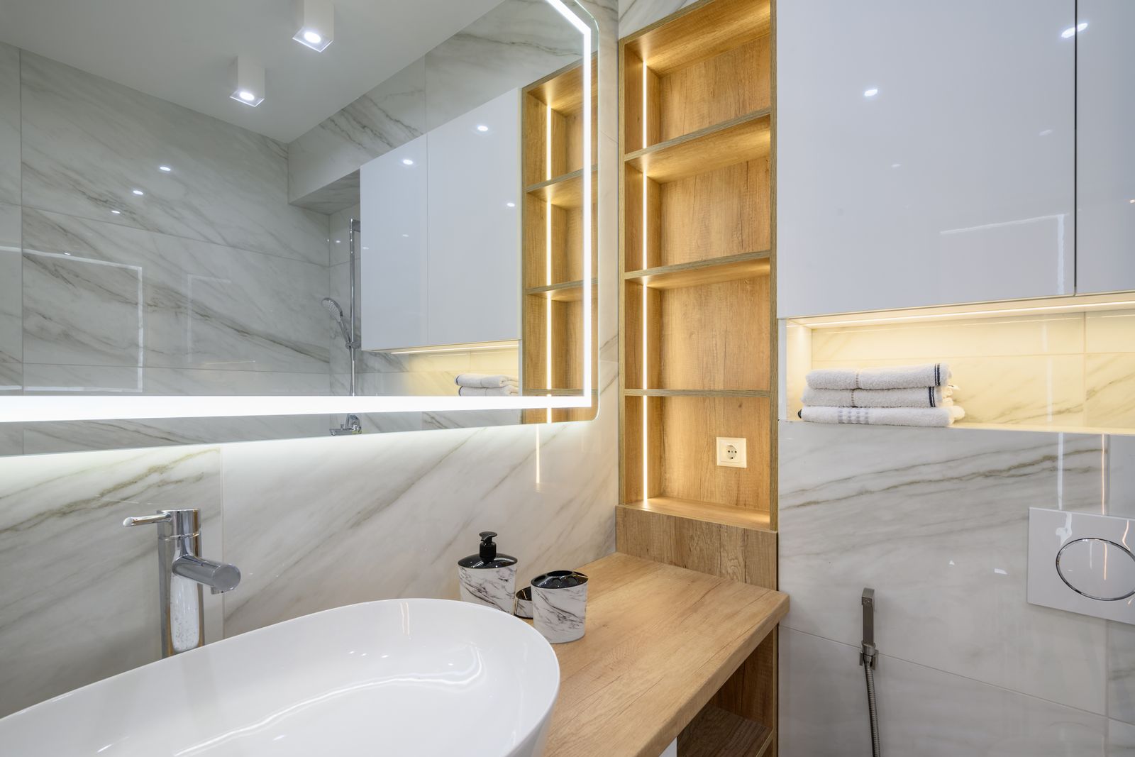 Badezimmer mit Marmorfliesen, einem weißen Aufsatzwaschbecken auf einer Holzplatte, einem großen Spiegel mit LED-Lichtband, eckigen Spots an der Decke und beleuchteten Holzregalen, daneben Handtücher in einer hell erleuchteten Nische unter weißen Hängeschränken