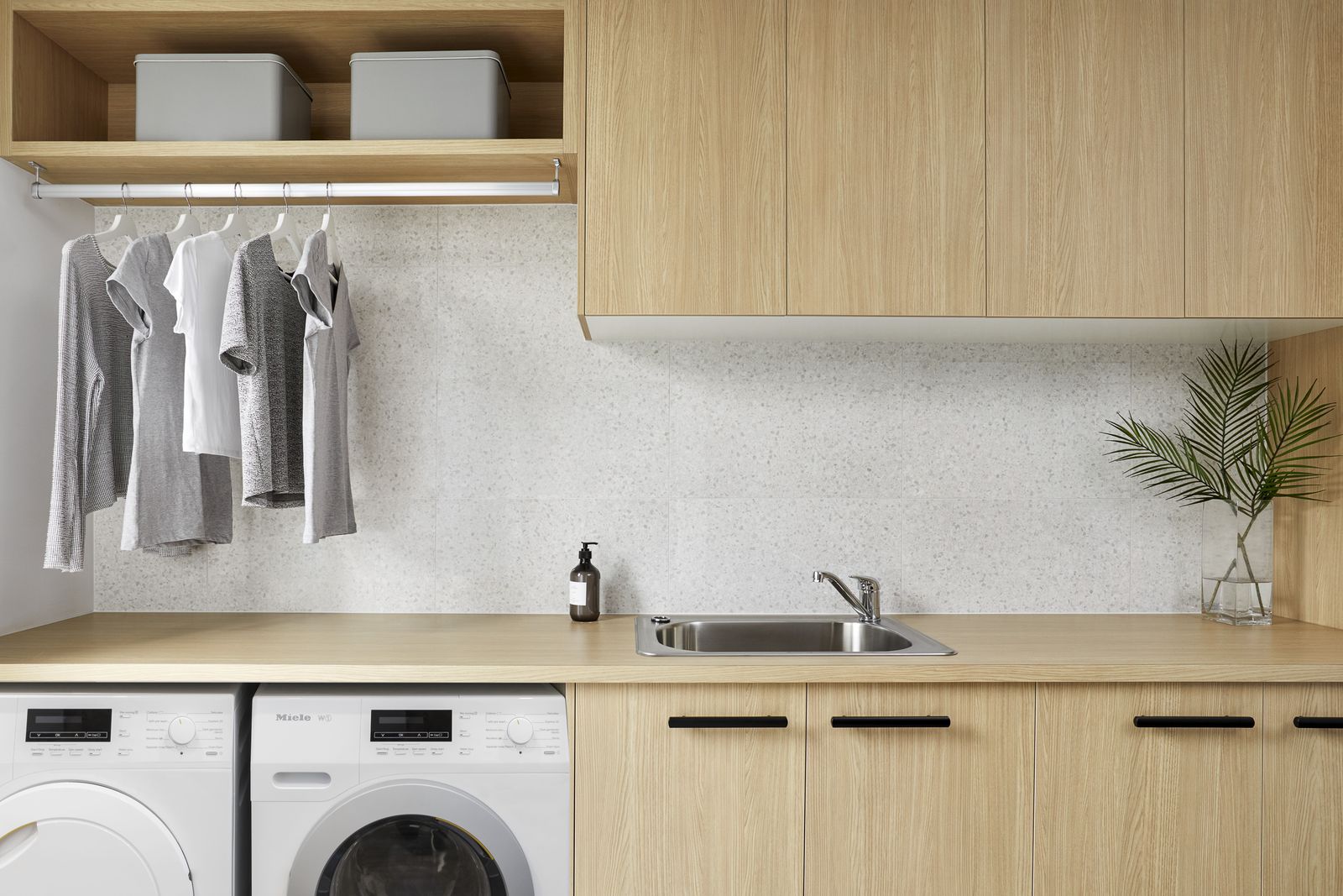 Hauswirtschaftsraum mit Waschmaschine und Trockner unter einer Arbeitsplatte aus Holz, Spülbecken, Holzschränken und einer Kleiderstange mit Shirts auf Bügeln