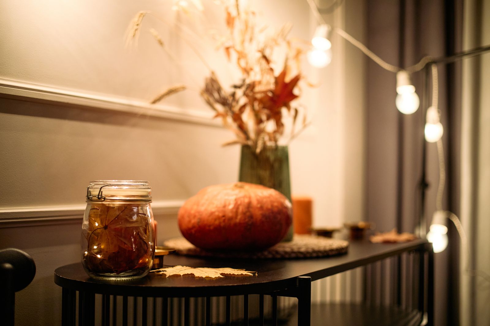 Kommode am Abend mit einem Kürbis, einem Glas mit Herbstlaub und Lichterkette, einer Vase mit Trockenblumen und Getreideähren, Kerzen und einer Lichterkette an der Wand
