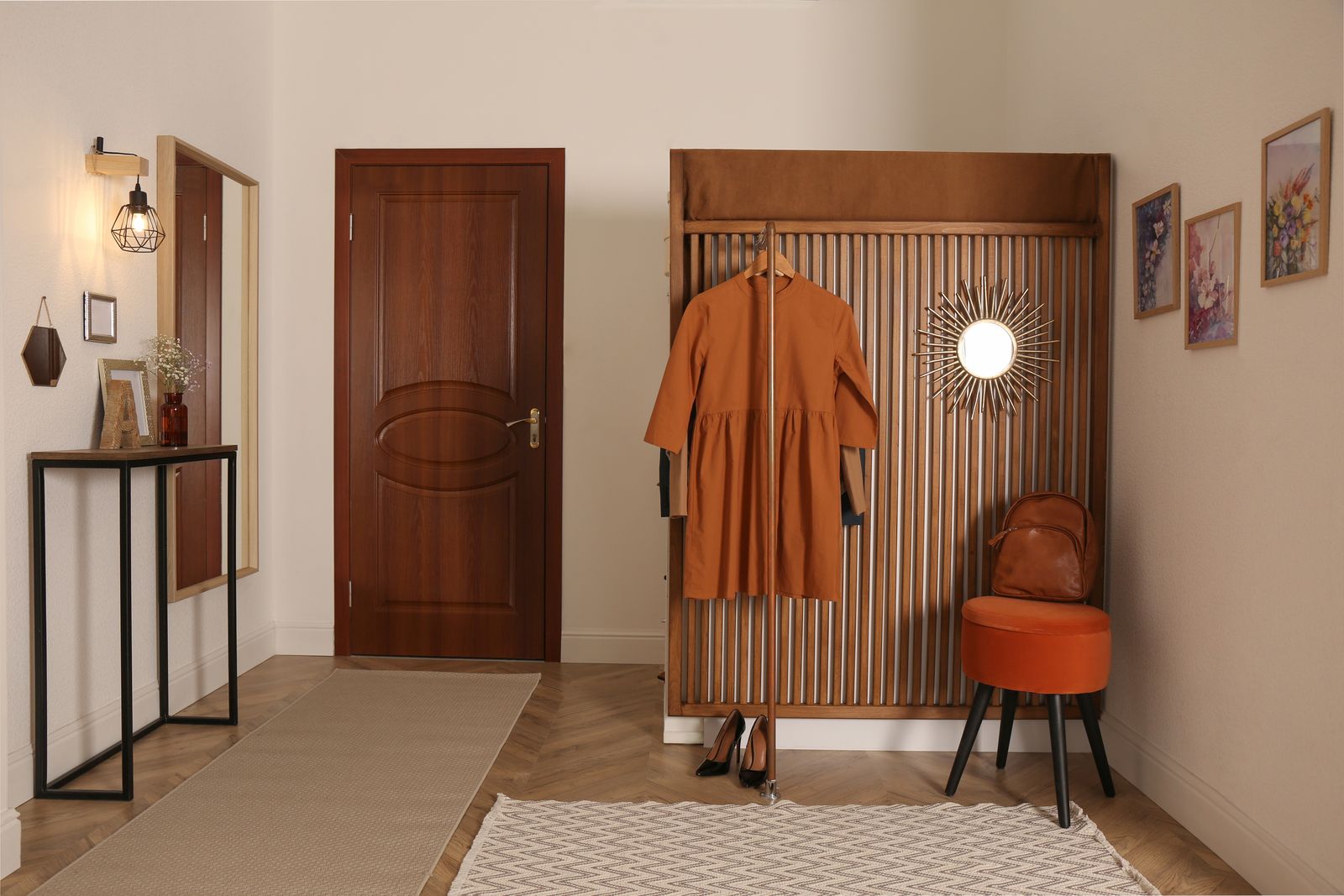 Warmer Eingangsbereich mit brauner Holztür, schmalem Konsolentisch, Wandleuchte, Lamellen-Garderobe mit rostrotem Kleid, Sonnenspiegel, orangefarbenem Hocker mit Rucksack und Läufern auf Fischgrätparkett