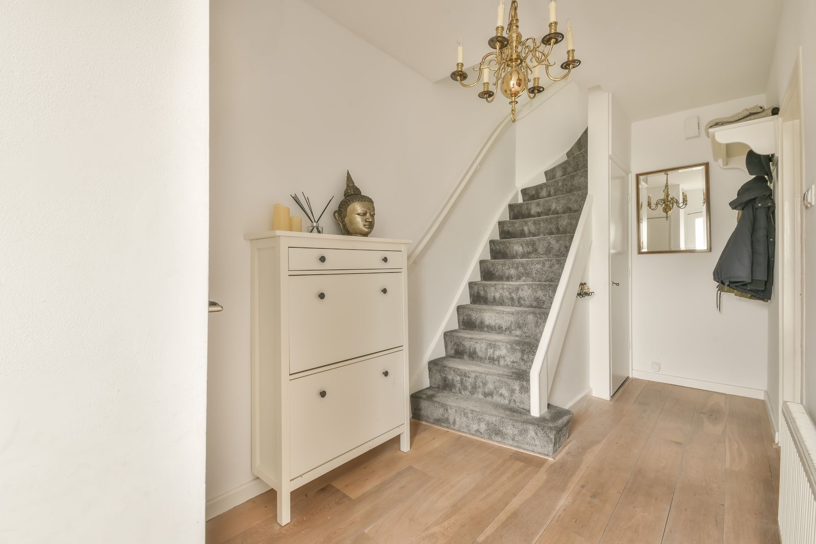 Heller Eingangsbereich mit grau bespannter Treppe, weißem Schuhschrank mit Kerzen und goldfarbener Buddha-Büste, Messing-Kronleuchter, Wandspiegel mit Goldrahmen, Jacke an der Garderobe und Holzdielen