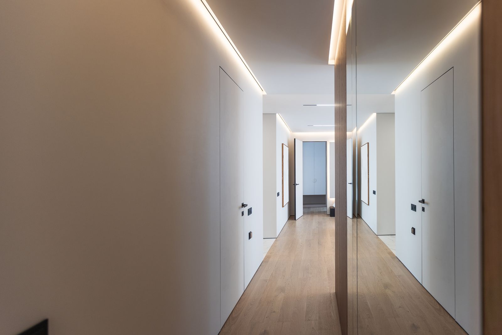Langer heller Flur mit indirektem Licht an der Decke, weißen Türen, Holzboden und einer Spiegelwand auf der rechten Seite