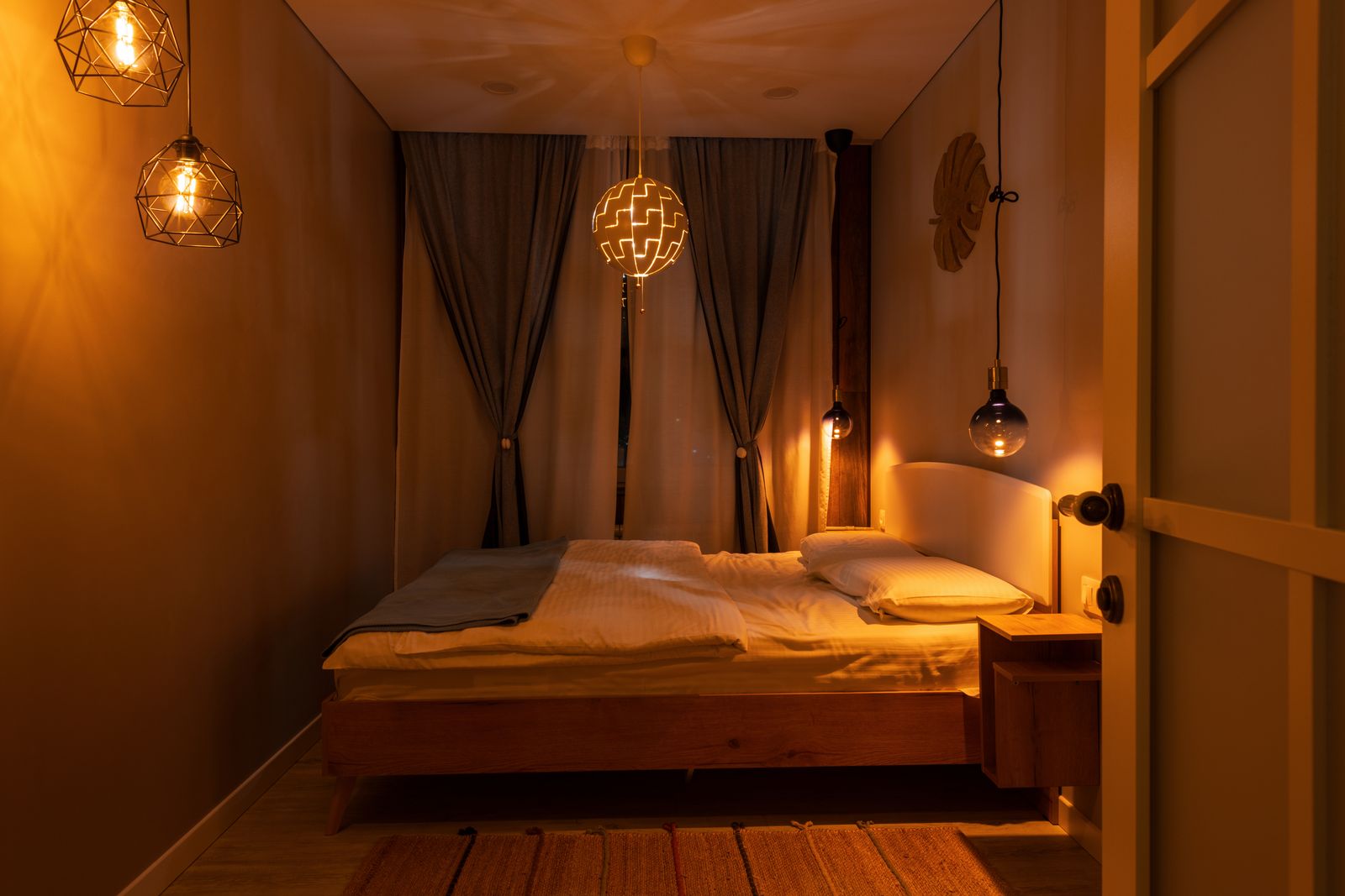 Schlafzimmer am Abend in warmem Licht mit Holzbett, mehreren Hängeleuchten aus Metallgitter und Glas, einer Kugelleuchte vor dem Fenster und einem Nachttisch