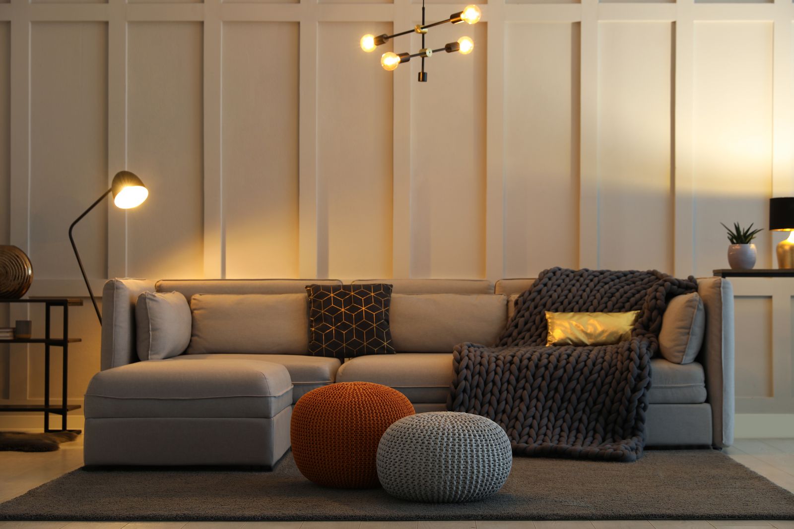Wohnzimmer am Abend mit hellem Ecksofa, grober Strickdecke und zwei Strickpoufs, beleuchtet von einer Stehlampe, einer Tischlampe und einer Hängeleuchte mit Glühbirnen