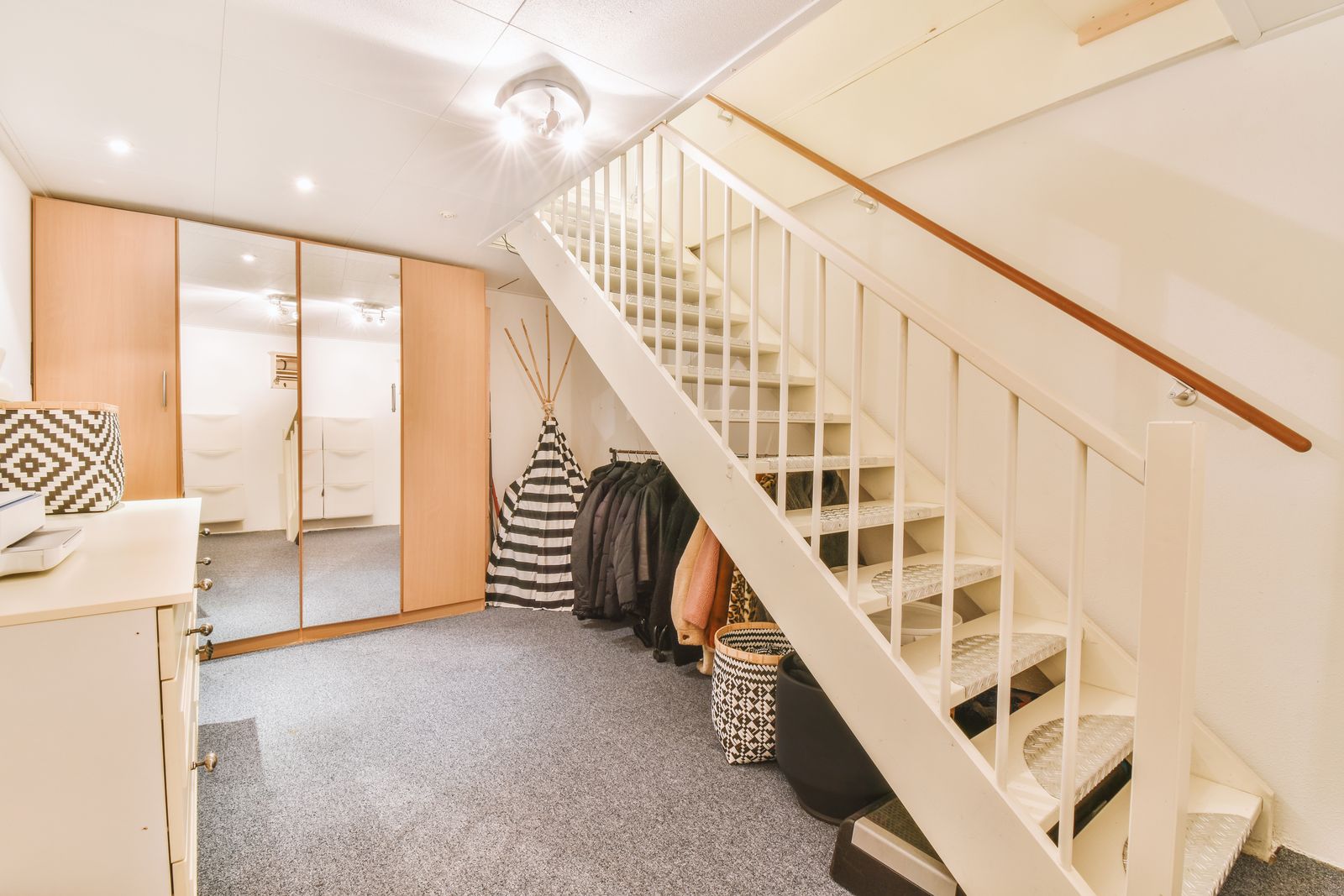 Ausgebauter Kellerraum mit heller Holztreppe, grauem Teppichboden, Kleiderschrank mit Spiegeltüren, Jacken an einer Garderobe unter der Treppe, Körben und einer weißen Kommode
