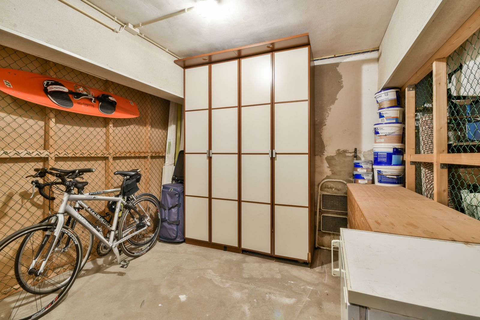 Kellerraum mit Betonboden, zwei Fahrrädern vor einer Lattenwand mit Maschendraht, einem Wakeboard an der Wand, einem hohen weißen Schrank, gestapelten Farbeimern und einem Holzregal