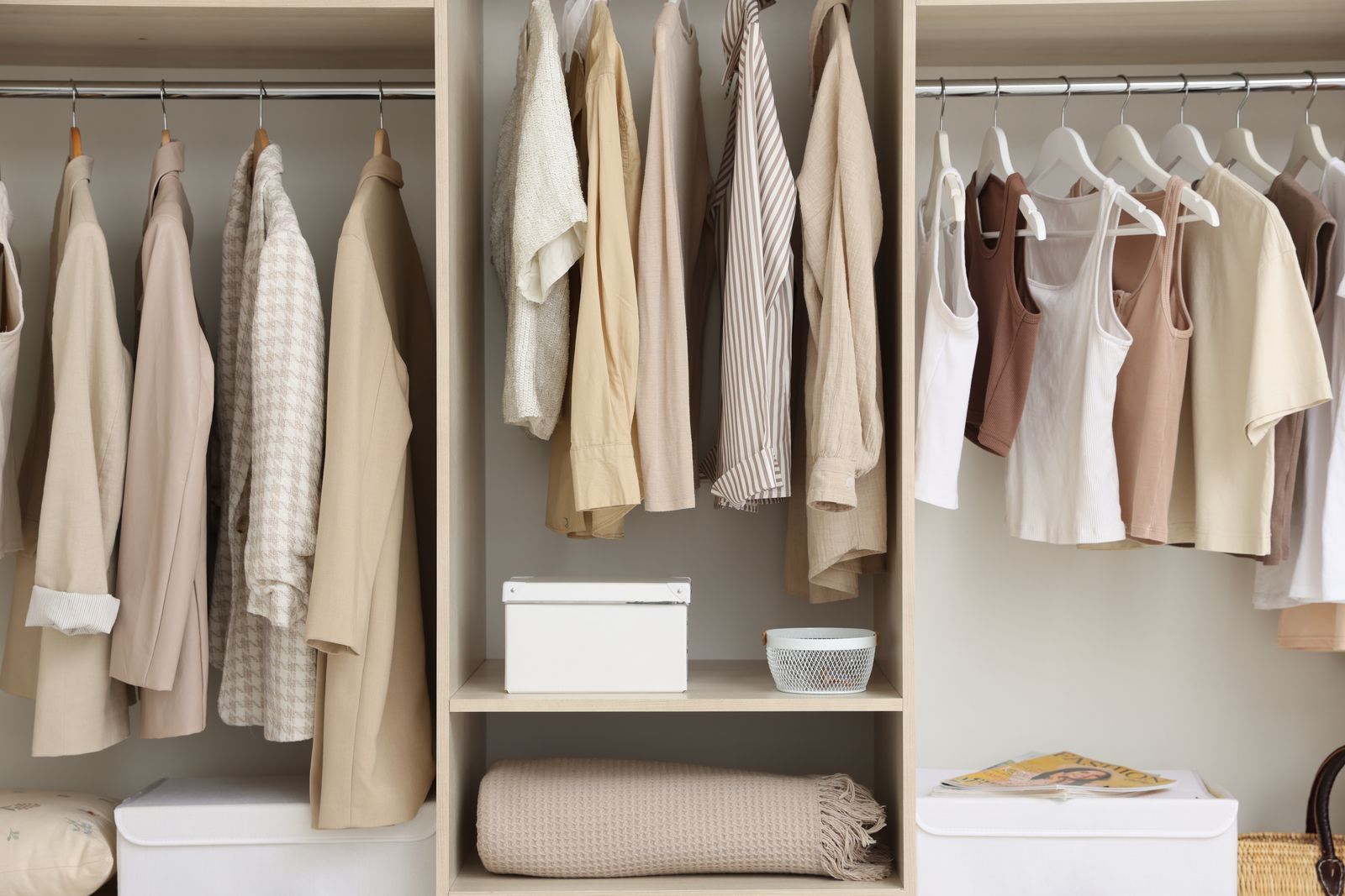 Geöffneter Kleiderschrank mit drei Fächern, Jacken und Oberteile in Beige- und Cremetönen an den Stangen, darunter ein Regalboden mit einer weißen Box, einem kleinen Korb und einer gefalteten Decke