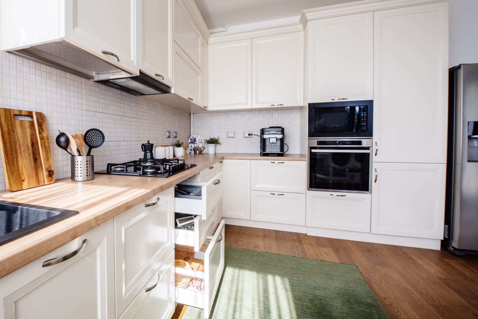 Helle Küche in L-Form mit cremeweißen Schränken, Holzarbeitsplatte, Gasherd und Schneidebrett, zwei geöffnete Schubladen neben der Spüle, Einbaubackofen, Kühlschrank und ein grüner Läufer auf dem Holzboden