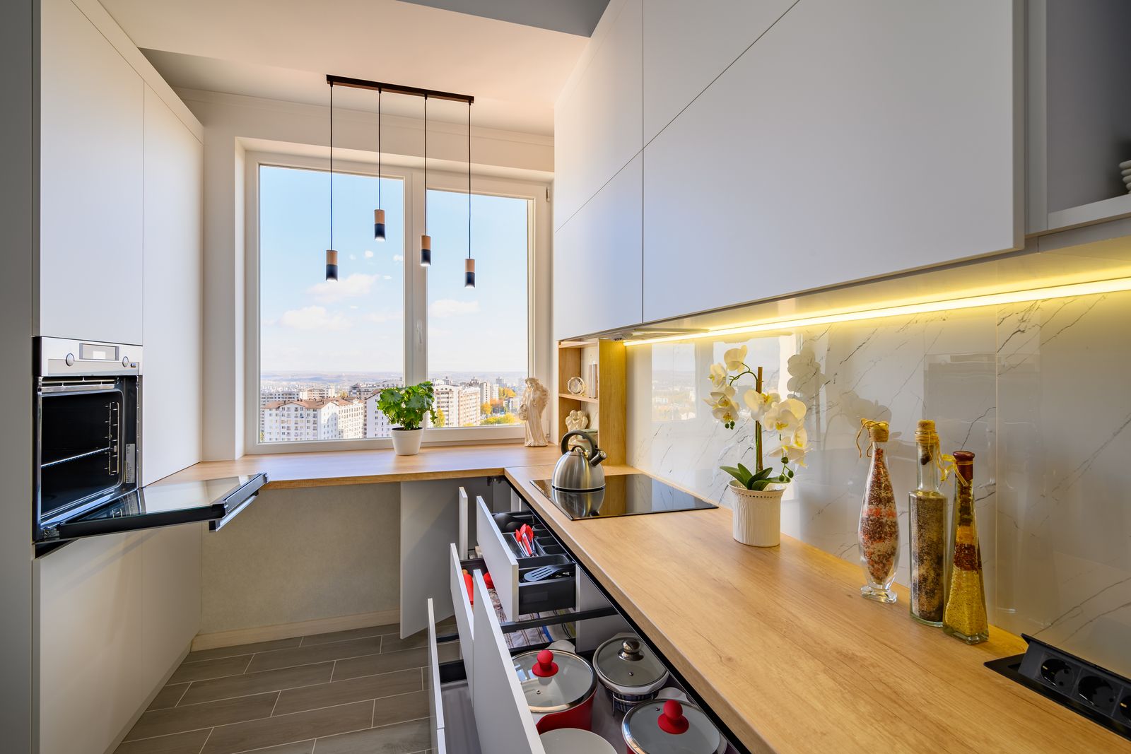 Kleine helle Küche in U-Form mit Fenster und Stadtblick, geöffnete Schubladen mit Besteckeinsatz und Töpfen, Holzarbeitsplatte mit Wasserkocher, Orchidee und Ölflaschen, darüber grifflose weiße Hängeschränke