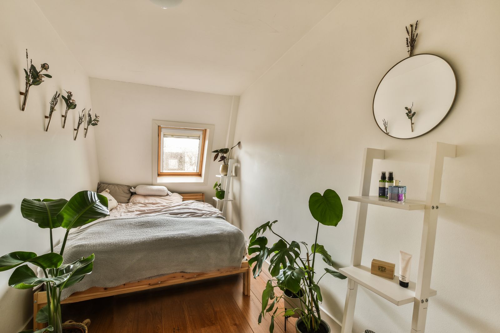 Kleines helles Schlafzimmer unter der Dachschräge mit Holzbett, grauer Strickdecke, Dachfenster, großen Pflanzen, rundem Spiegel und weißem Leiterregal