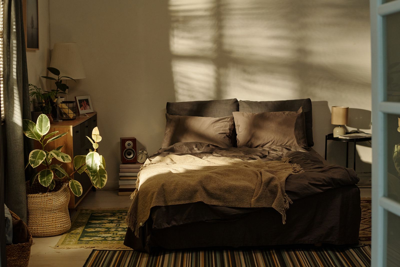 Schlafzimmer im warmen Abendlicht mit Doppelbett, grauer Bettwäsche und Wolldecke, daneben ein Nachttisch mit Lampe, eine Holzkommode, Pflanzen und ein Korb