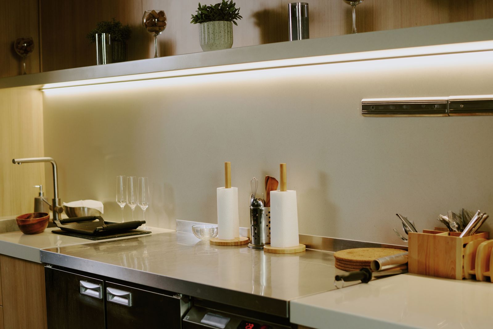 Küchenzeile mit Edelstahl-Arbeitsplatte und Spüle, darüber ein offenes Holzregal mit Pflanzen und Gläsern und einem LED-Lichtband darunter, auf der Arbeitsplatte Küchenrollenhalter, Sektgläser und ein Besteckkasten aus Holz