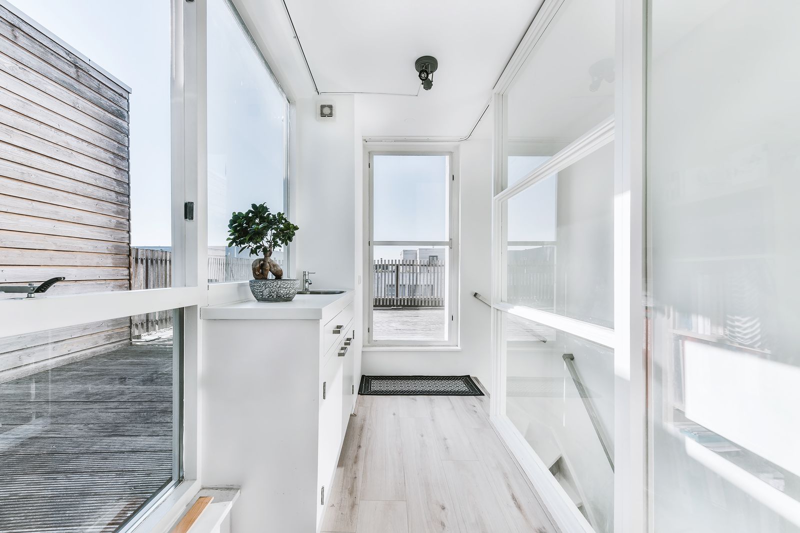 Heller schmaler Gang mit großen Fenstern zur Dachterrasse, einem weißen Unterschrank mit Bonsai, hellem Holzboden, einer Fußmatte und einer Glastür am Ende
