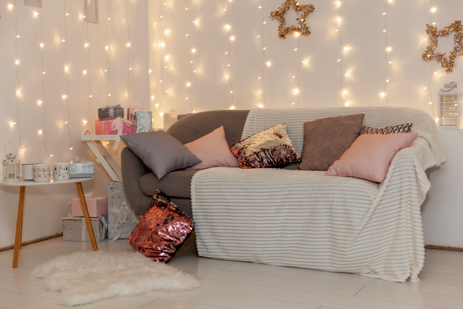 Graues Sofa mit heller Kuscheldecke und rosa, grauen und Pailletten-Kissen vor einer Wand mit einem warmweißen Lichtervorhang und goldenen Sternen, daneben ein Beistelltisch mit Tassen, Geschenke, Laternen und ein Schaffell auf dem Boden