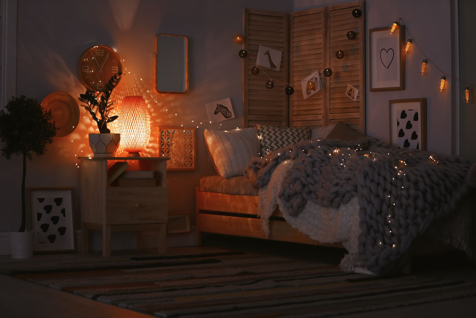 Bett mit grober Strickdecke und Lichterkette im Dunkeln, daneben ein Nachttisch mit leuchtender Rattanlampe und Pflanze, Lichterketten an Wand und Paravent, Bilderrahmen, Wanduhr und ein gestreifter Teppich