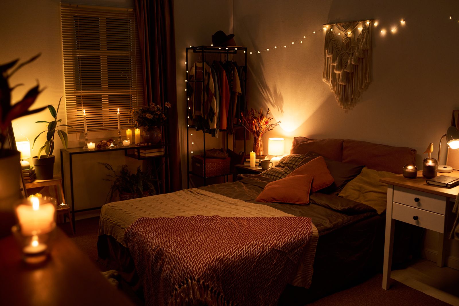 Schlafzimmer am Abend mit Doppelbett, Kissen und Strickdecke, Lichterkette und Makramee an der Wand, Nachttisch mit Leselampe und Windlichtern, Kerzen auf einer Konsole am Fenster und einer kleinen Tischlampe neben einem Kleiderständer