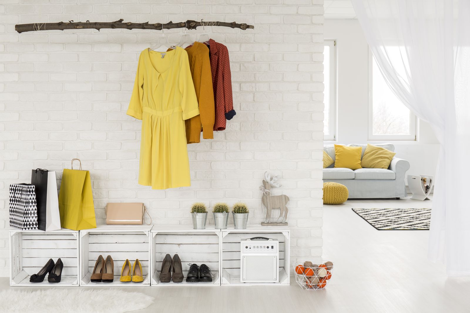 Weiße Backsteinwand mit Kleidern an einem Ast als Garderobe, darunter weiße Holzkisten als Schuhregal mit Pumps und Schnürschuhen, rechts der Blick ins Wohnzimmer