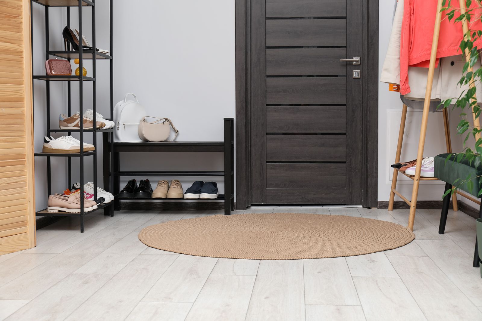 Eingangsbereich mit dunkler Holztür, schwarzem Metallregal voller Schuhe und Taschen, einer Schuhbank mit vier Paar Schuhen und einem runden Teppich auf hellem Boden