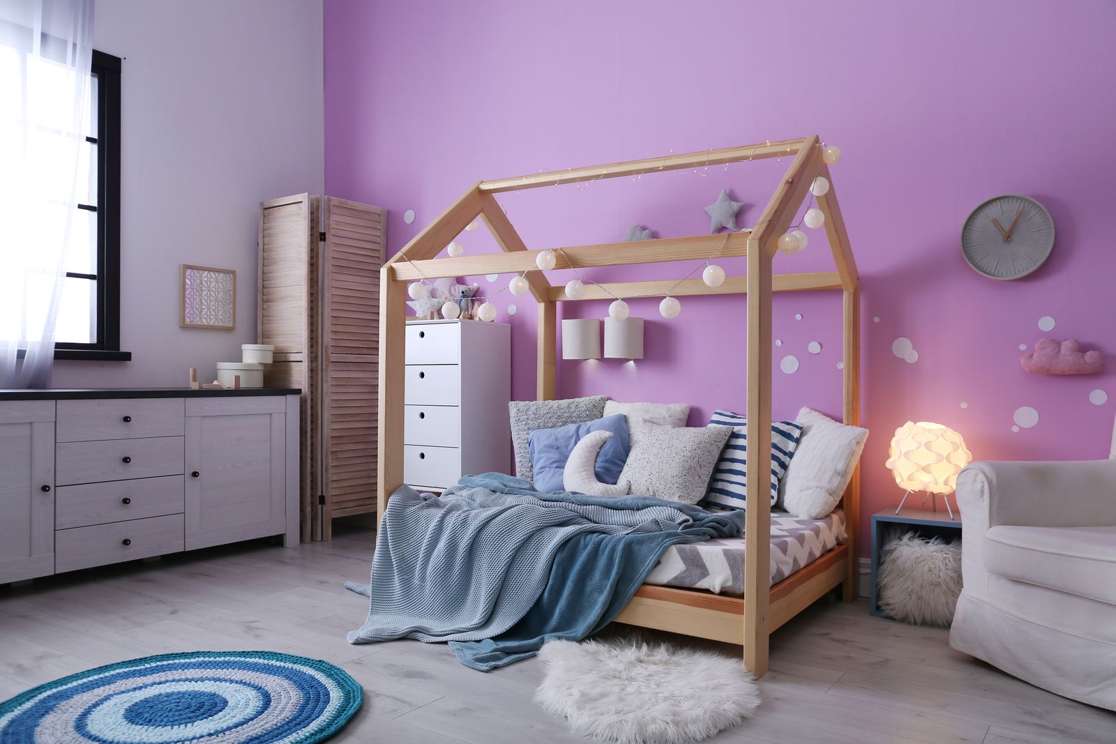 Kinderzimmer mit fliederfarbener Wand, Hausbett aus Holz mit Lichterkette, Kissen und blauer Decke, daneben eine weiße Kommode, ein Sideboard, ein Sessel und ein runder blauer Häkelteppich