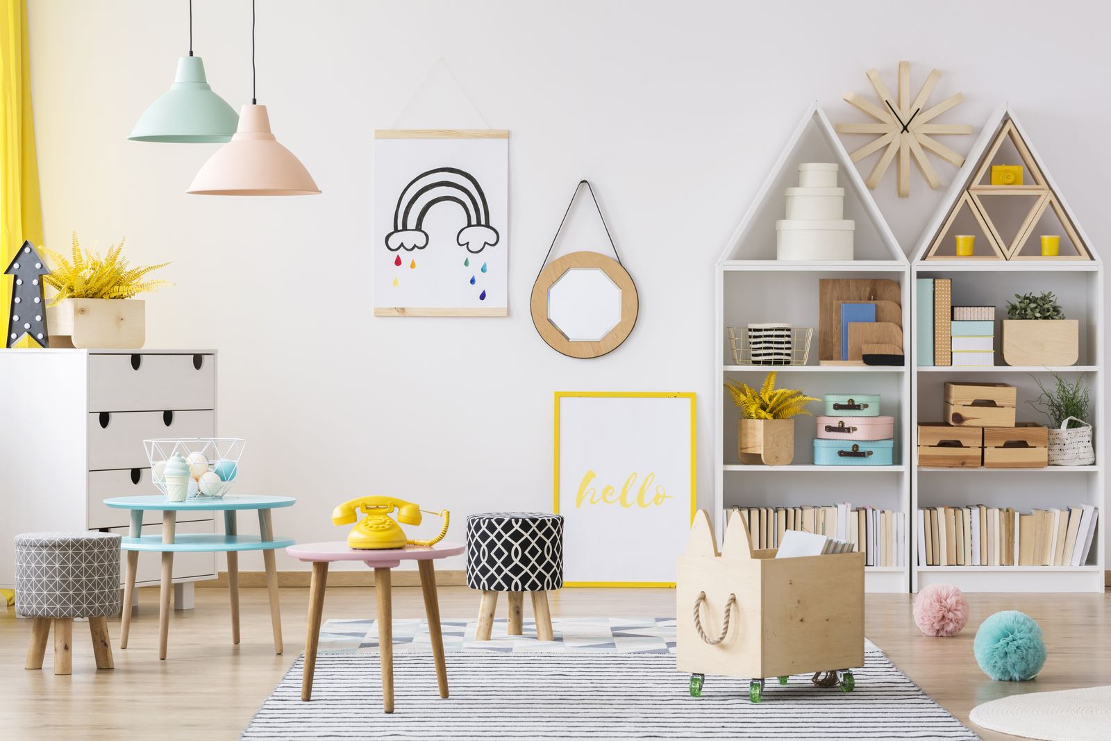 Helles Spielzimmer mit zwei weißen Regalen in Hausform voller Bücher, Holzkisten und bunter Schachteln, davor eine Holzkiste auf Rollen, ein kleiner Tisch mit gelbem Telefon, Hocker und ein gestreifter Teppich